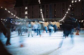Skaters on an outdoor ice rink after dark, lit by strings of warm white bulbs overhead