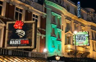 Illuminated theater marquees for Hadestown at the Apollo Theatre and Faulty Towers The Dining Experience in London's West End at night, with glowing streetlights and people walking below.