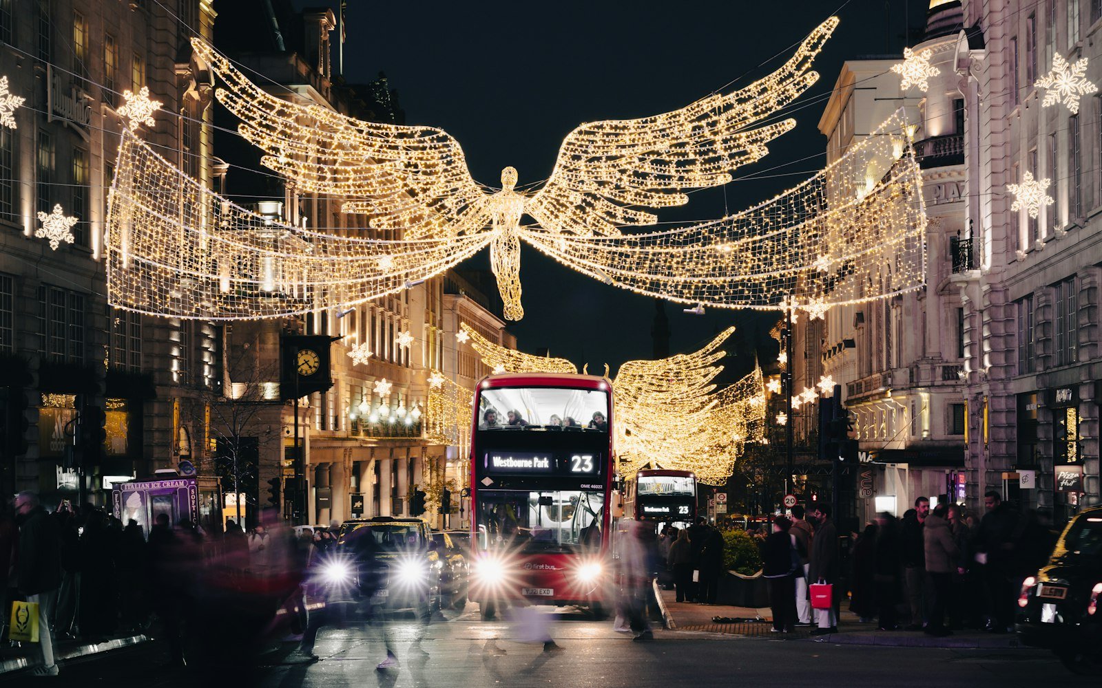 A London street at night lined with large illuminated angel decorations suspended above the road, with buses and shoppers below