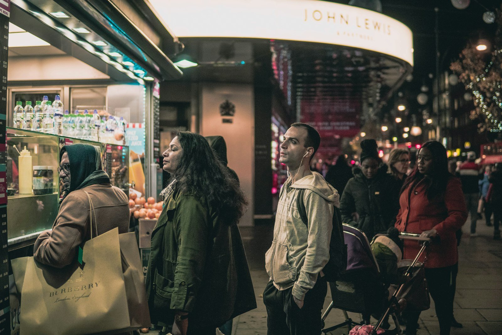 Shoppers queuing in a line along the pavement beside a brightly lit London store after dark