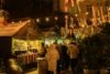 Visitors gathered around lit wooden stalls at an outdoor Christmas market after dark, holding hot drinks