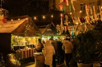 Visitors gathered around lit wooden stalls at an outdoor Christmas market after dark, holding hot drinks