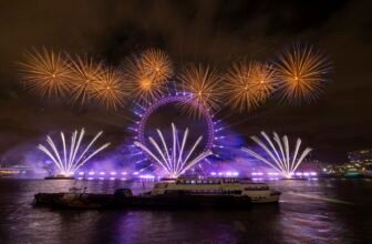 Fireworks bursting in gold and white above the floodlit London Eye at night, reflected in the River Thames